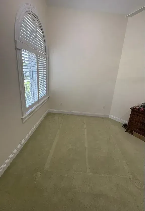 Empty beige bedroom with green carpet, white blinds, and a small dresser in the corner