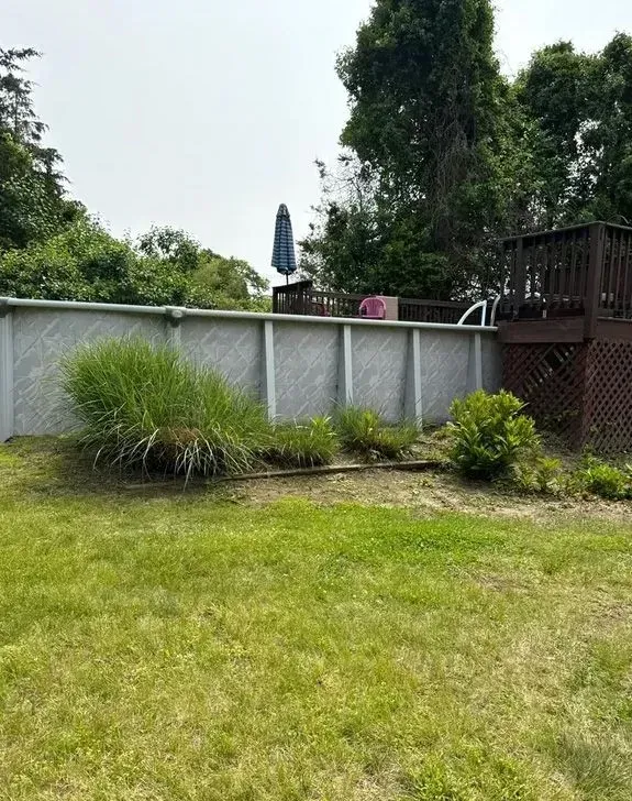 Backyard lawn with shrubs, a white fence, and tall trees under a partly cloudy sky