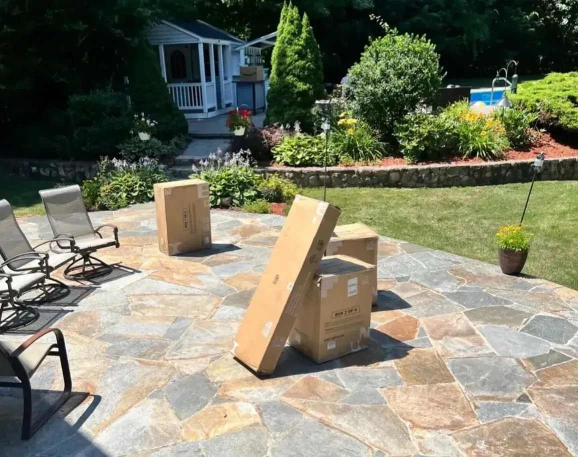 Backyard patio with stacked cardboard boxes and a slanted board on stone paving, near chairs and a garden bed