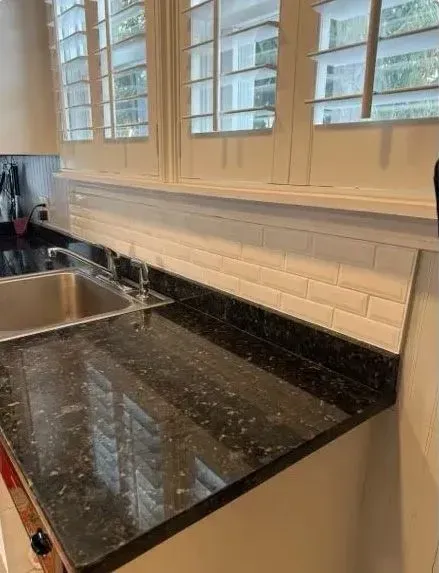 Kitchen countertop with sink, black granite surface, white subway tile backsplash, and window shutters above.