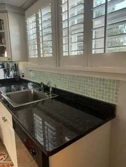 Modern kitchen counter with black granite, double sink, and white shutters above a green tile backsplash