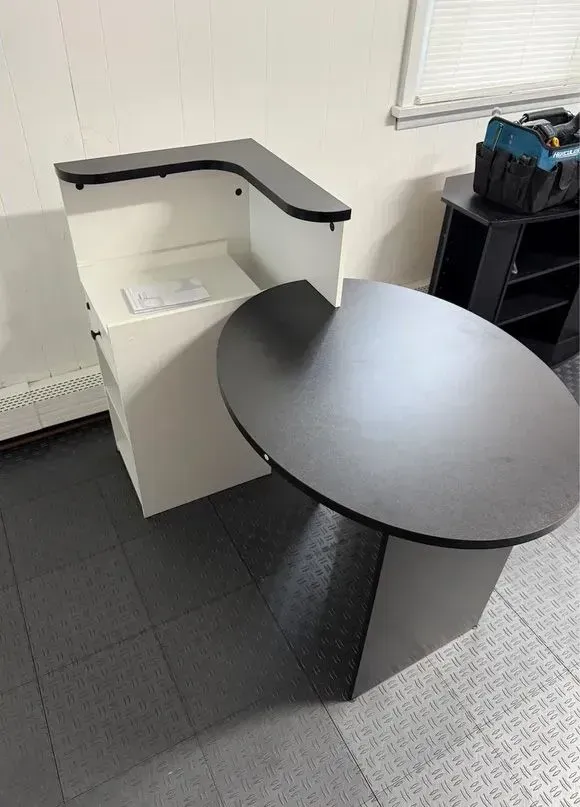 Small office desk with a black oval tabletop and white partition, next to a window and radiator.