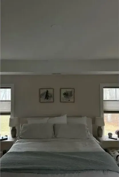 Neatly made bed in a bright bedroom with two windows and framed wall art above the headboard