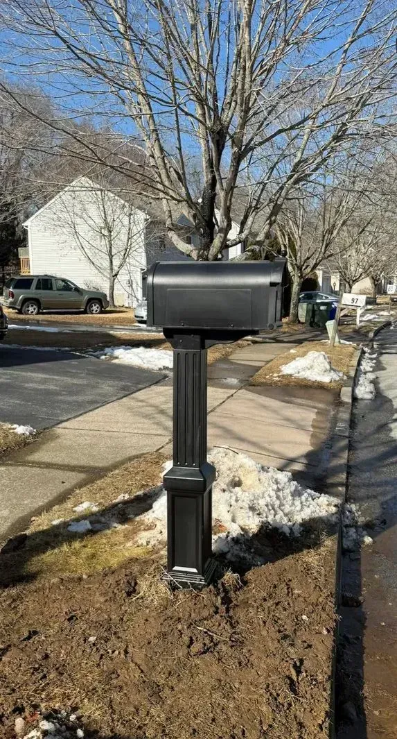 Black mailbox on a post in a snowy suburban yard with trees and houses behind it