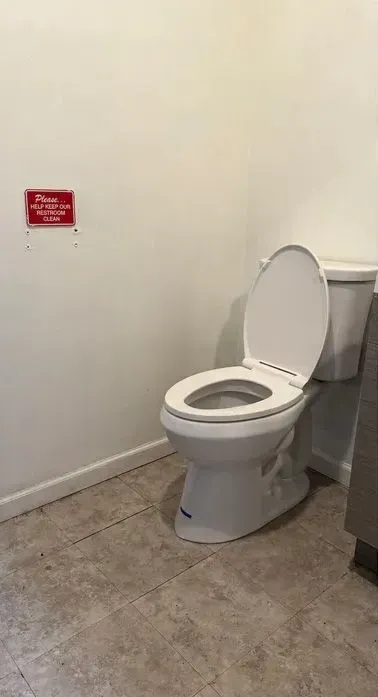 Small restroom with a toilet, white walls, gray tile floor, and a red sign on the wall