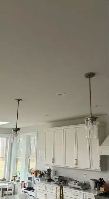 Modern white kitchen with pendant lights, cabinets, and a countertop by tall windows.
