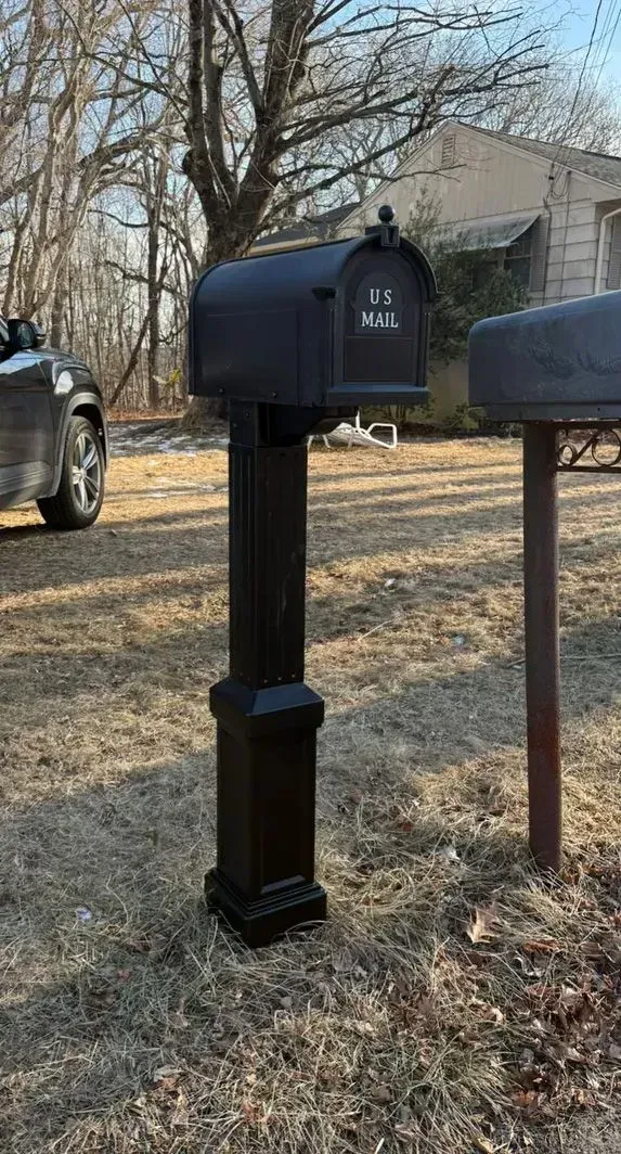 Black mailbox on a post beside another mailbox in a grassy yard with trees and a house in the background