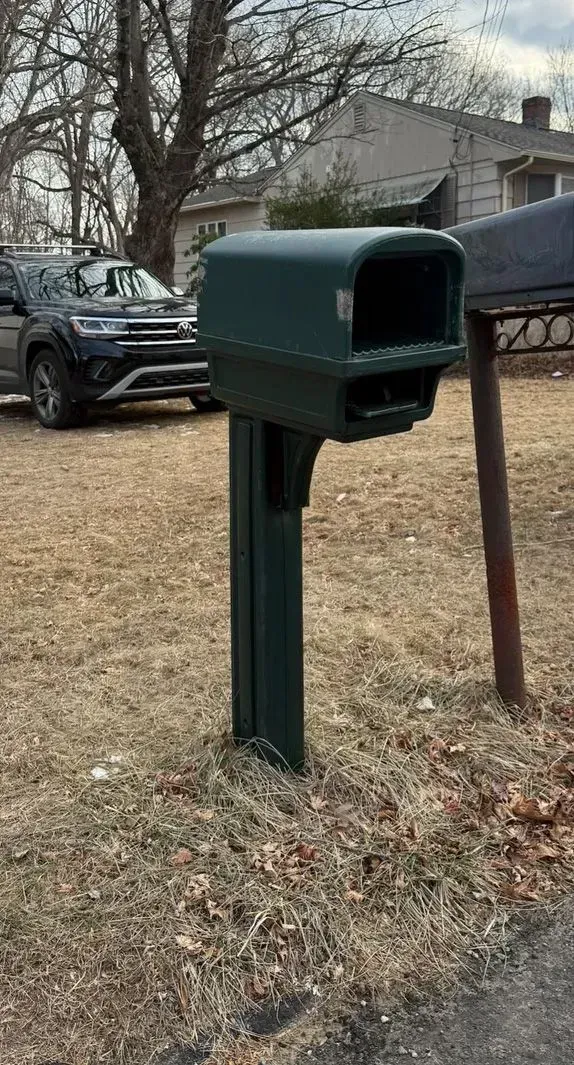 Green mailbox on a post in a yard, with a house and parked vehicles in the background.