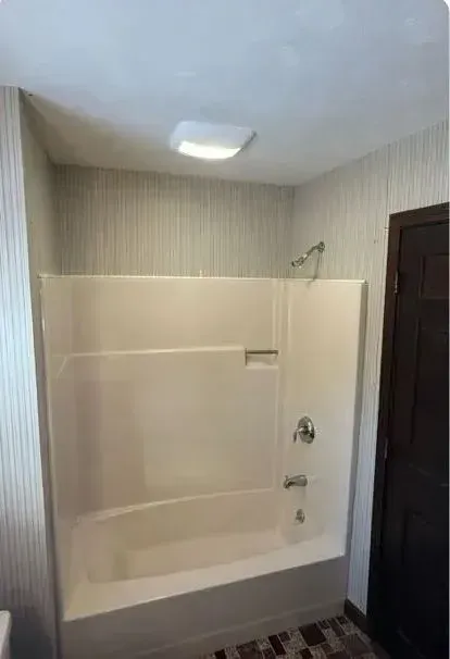 Bathroom with a white bathtub-shower combo, beige striped walls, ceiling light, and a dark wood door.