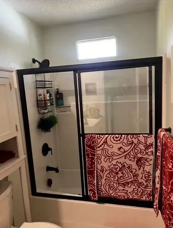 Bathroom with a black-framed tub/shower enclosure, red patterned curtain, and small window above.