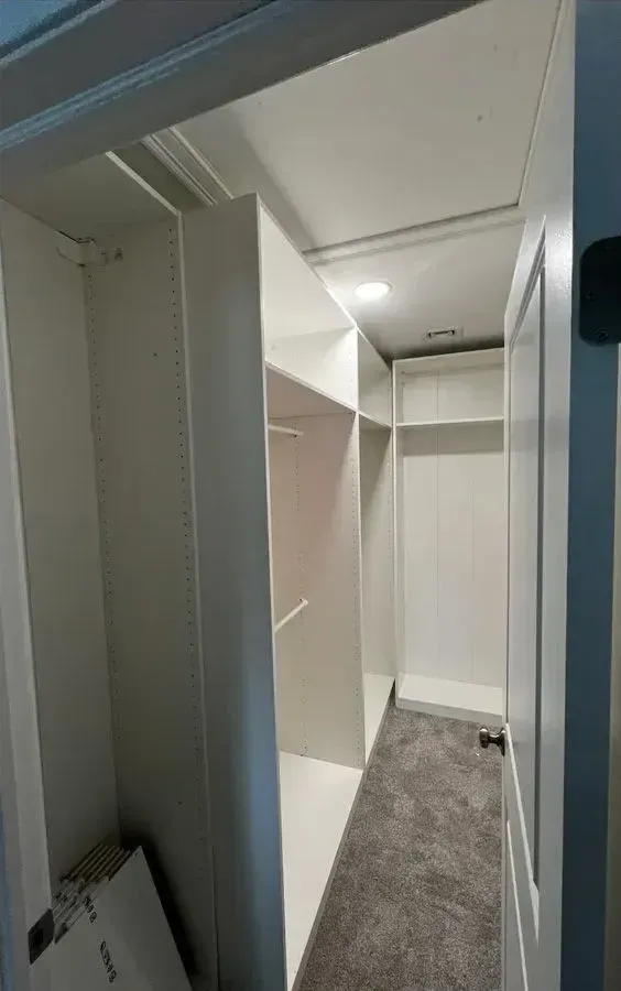 Empty walk-in closet with white built-in shelves and hanging rods, gray carpet, and a ceiling light.