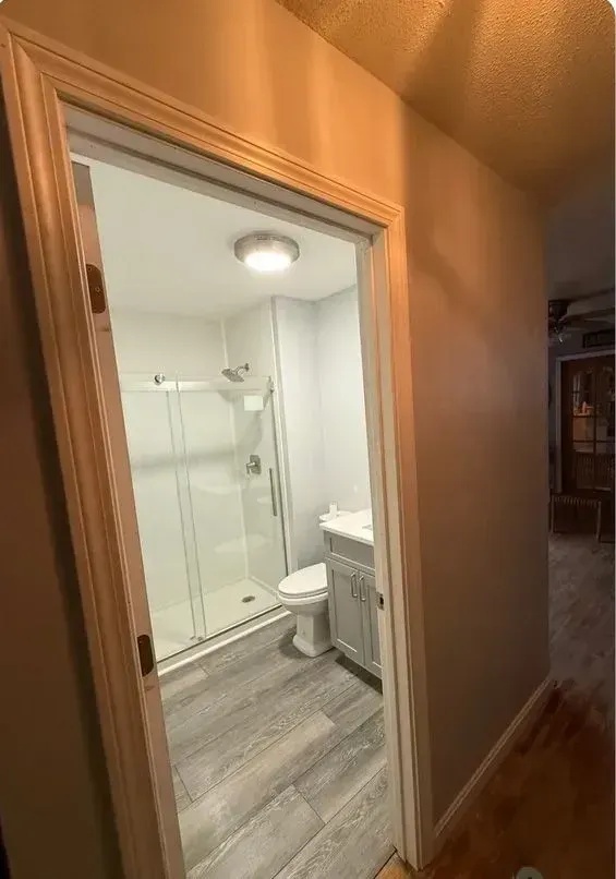 Hallway view into a bright bathroom with glass shower, toilet, vanity, and wood-look flooring.