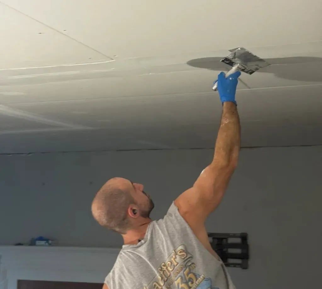 Man scraping joint compound from a ceiling seam with a trowel, wearing a blue glove