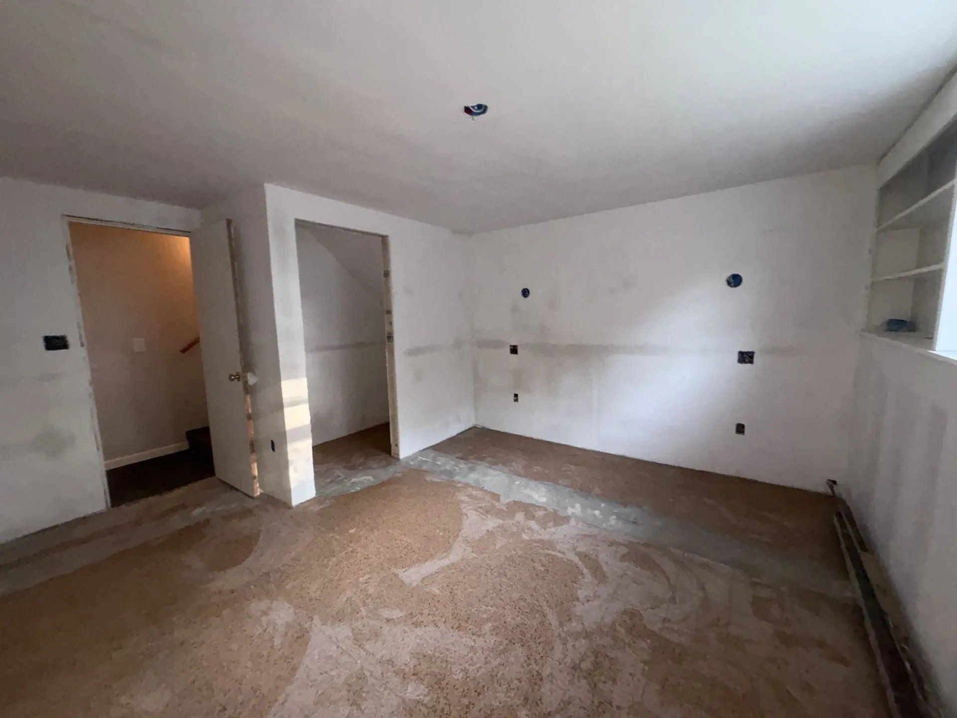Empty white room under renovation with unfinished floor, wall outlets, and open doorway