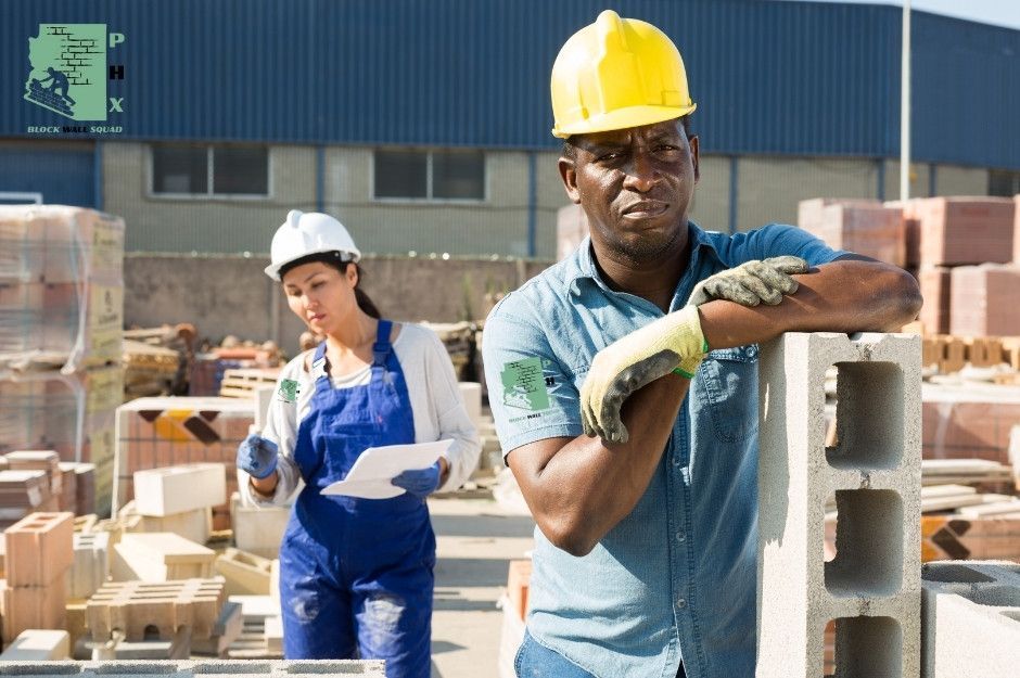 Block wall repair companies work on a commercial masonry project, with the lead worker in a yellow hard hat leaning on a block. Stacks of concrete blocks are organized throughout the construction site at Cll Sahuaro, and E Calle Barbarita in Guadalupe, AZ.