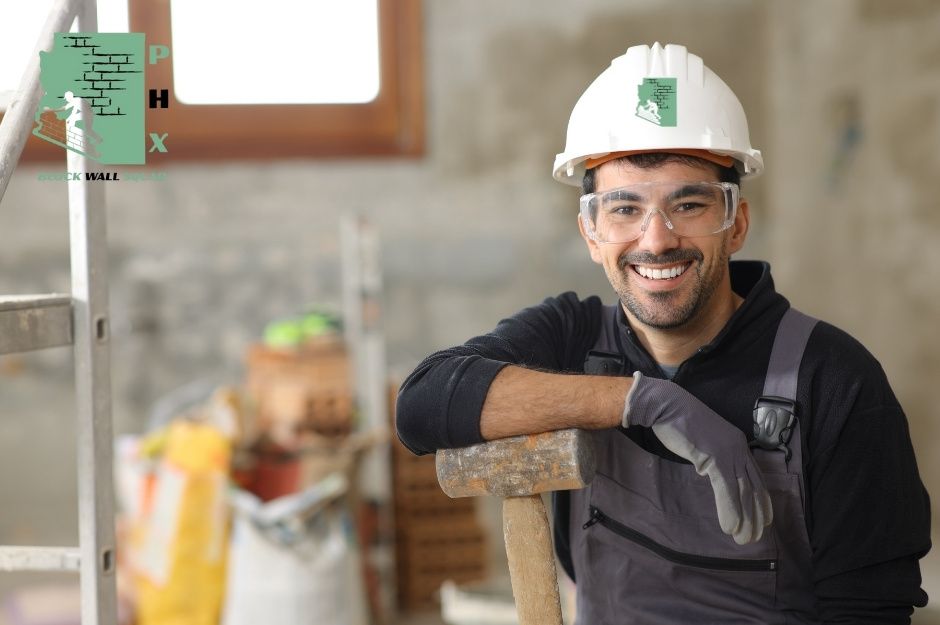 A smiling worker from a block wall repair company wearing a white hard hat and dark work shirt leans on a wooden-handled sledgehammer. He has safety glasses and appears to be on a commercial construction site with concrete block walls visible in the background near Saguaro Dr and Ocotillo Ln, Black Canyon City, AZ.