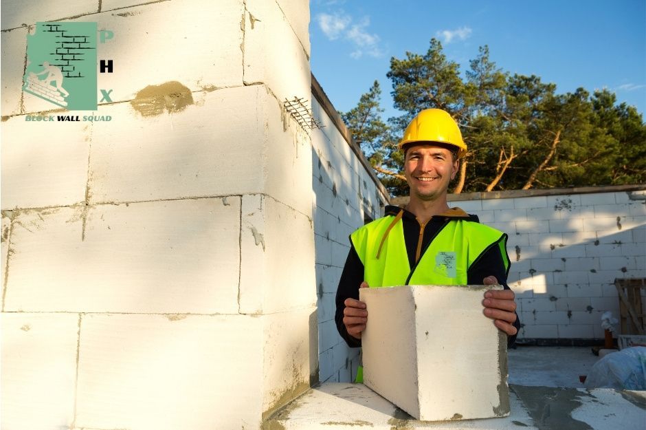A block wall repair company worker wearing a yellow hard hat and bright green safety vest holds white concrete blocks while standing next to a partially constructed residential block wall project. Blue sky and trees are visible in the background near N Spur Cross Rd, and Maguay Rd, Cave Creek, AZ.