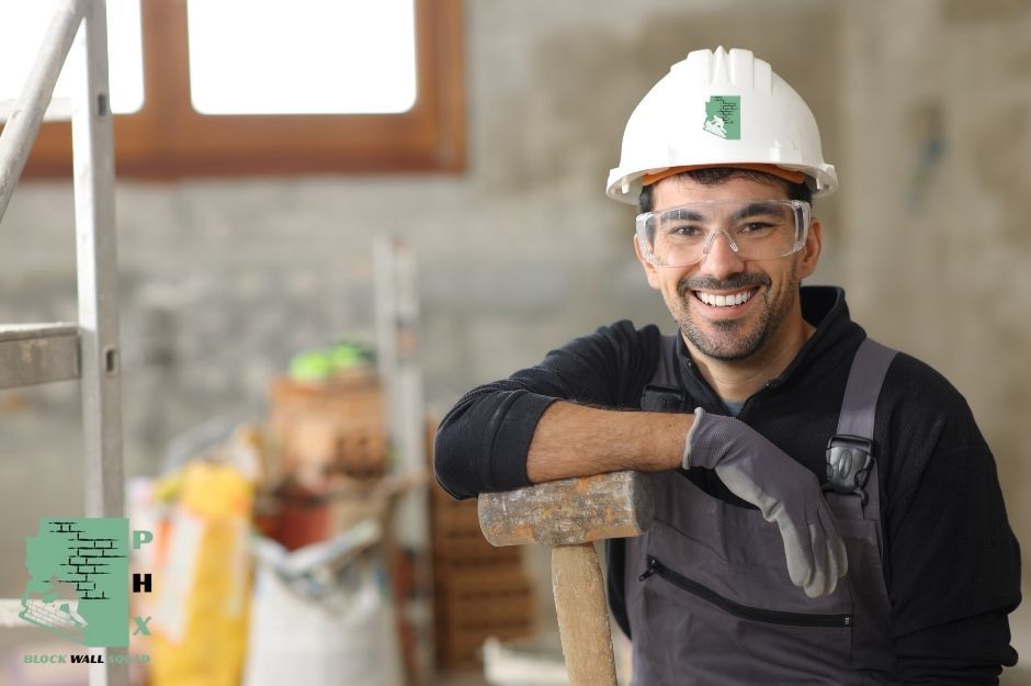 A smiling block wall repair company worker wearing a white hard hat and safety glasses leans on a wooden-handled sledgehammer at a residential construction site. He is dressed in dark work clothes with tool belts, standing among scaffolding and building materials near W Chandler Blvd, and N Kyrene Rd in Chandler, AZ.