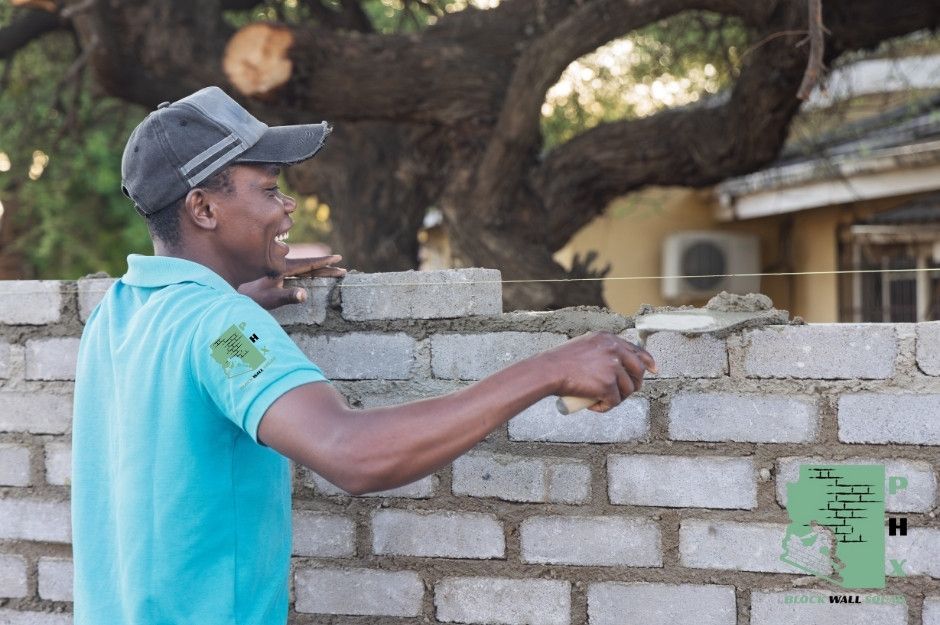A block wall repair company worker in a light blue shirt and cap performs residential masonry work, using a trowel to apply mortar between concrete blocks. A residential home is visible in the background surrounded by greenery near S 55th Dr, and S 55th Ave in Laveen, AZ.