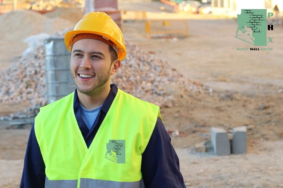 A smiling block wall repair company worker wearing an orange hard hat and bright yellow safety vest stands confidently at a commercial construction site. Behind him are piles of gravel and concrete blocks ready for the masonry project near N 9th St and Speer Trail New River, AZ.