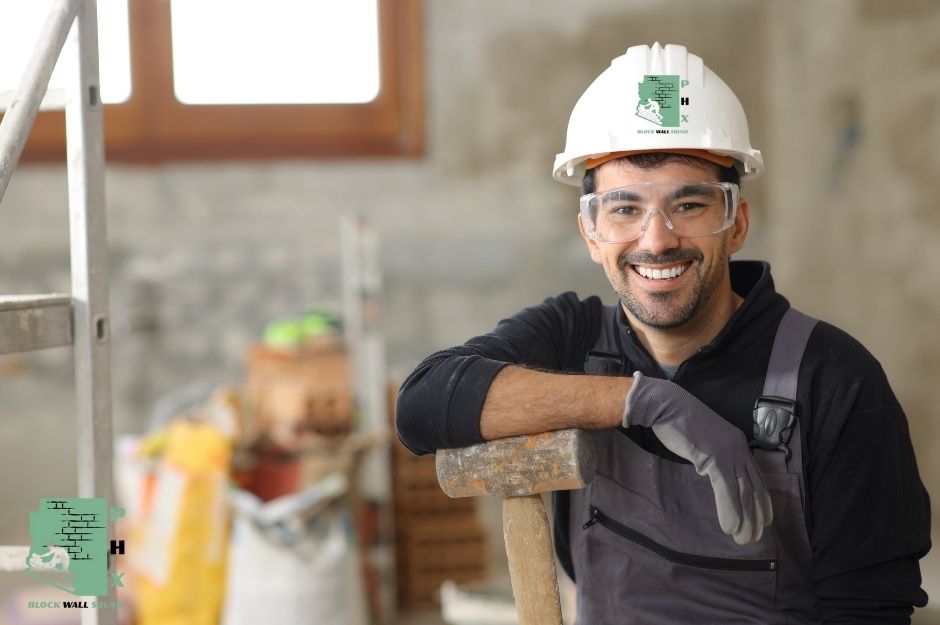 A block wall repair company worker in a white hard hat and safety glasses smiles while leaning on a hammer at a commercial site with visible concrete blocks near E Clairidge Dr, and N Candlewood Dr in Johnson Ranch, San Tan Valley, AZ.
