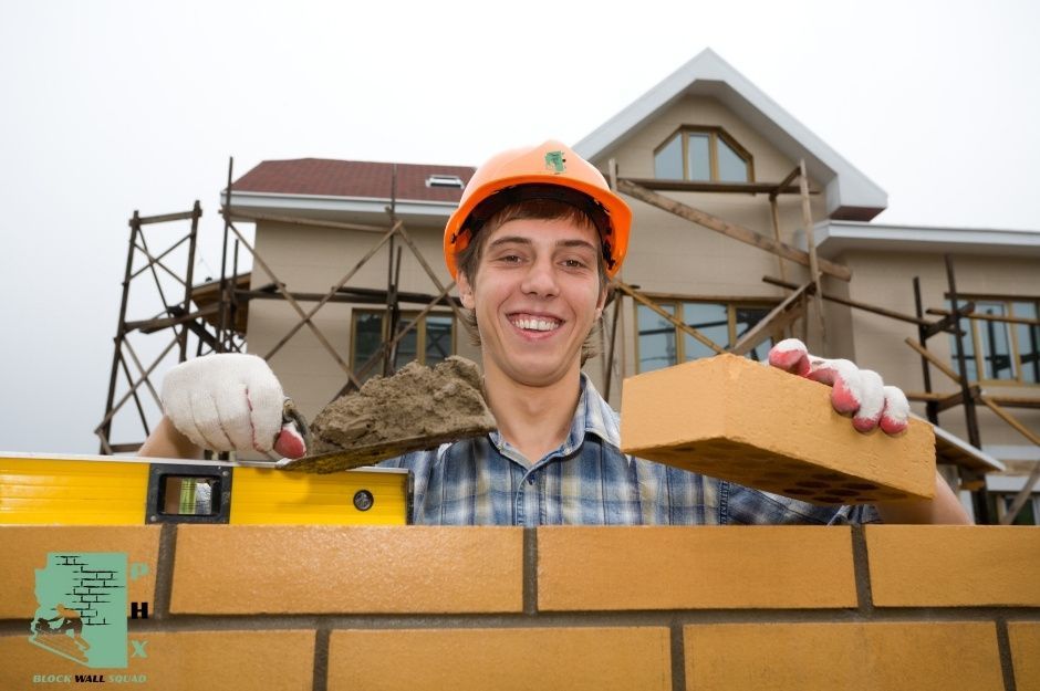 A block wall repair contractor wearing an orange hard hat and plaid shirt smiles while holding a brick and a trowel. He stands behind a partially built brick wall with a house under residential construction visible in the background near S Butte St, and E Third Aly, Dewey–Humboldt, AZ.