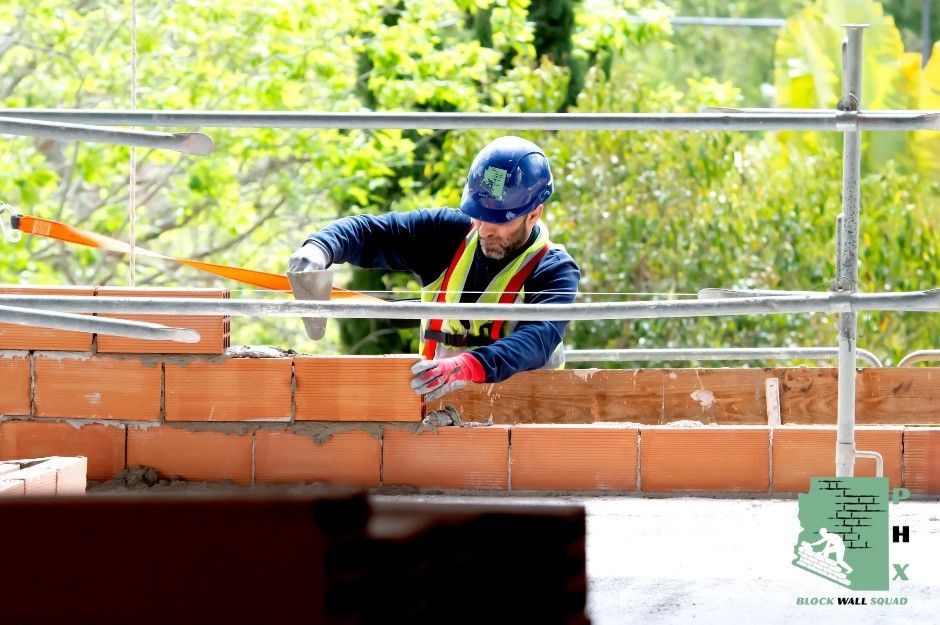 A block wall repair contractor wearing a blue hard hat is using a trowel to work on a red brick wall. Metal railings are visible above, with green foliage in a residential project. The worker is performing professional masonry work at W Cactus Wren Way and W Spirit Way in Florence, AZ.
