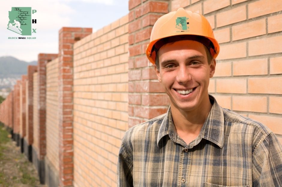 Block wall repair contractor smiling while wearing an orange hard hat and plaid shirt, standing in front of a commercial brick block wall under construction with sections visible extending into the distance. The worker appears confident and professional, positioned against the backdrop of the masonry work. Located near E Woodman Dr and Rural Rd, Tempe, AZ.