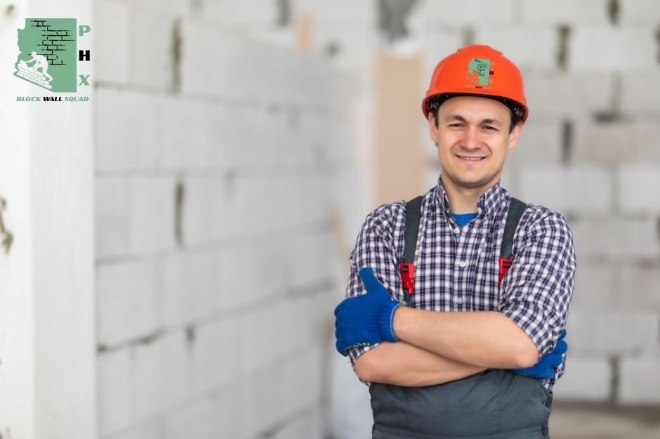 A professional block wall repair handyman with an orange hard hat and a gray work apron smiles with his arms crossed at a residential site, with concrete block walls behind him, near E Rosa Rd, and 22nd St in Charleston Estates, Queen Creek, AZ.
