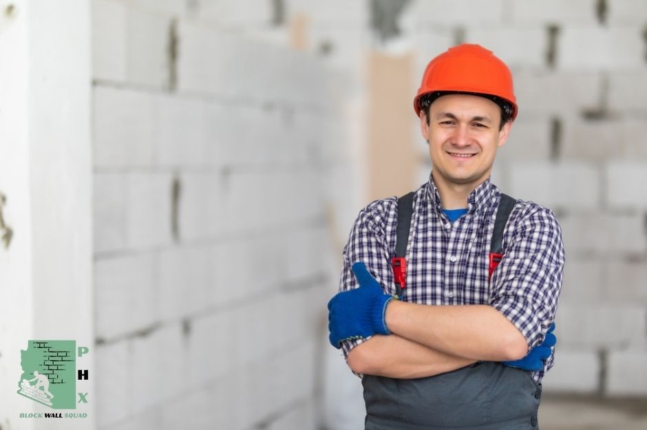 A confident block wall repair specialist wearing an orange hard hat, checkered shirt, and blue work gloves stands with arms crossed in front of a residential concrete block wall project. He is wearing overalls and has a friendly smile on his face near E Paint Pony, and Paint Pony Dr, Carefree, AZ.