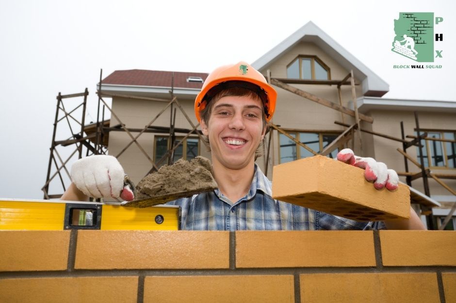 A confident block wall repair specialist in an orange hard hat and plaid shirt holds a masonry trowel while laying bricks on a wall. He is working on a residential project near W Lindner Ave, and S Johnson in Dobson Ranch, Mesa, AZ.
