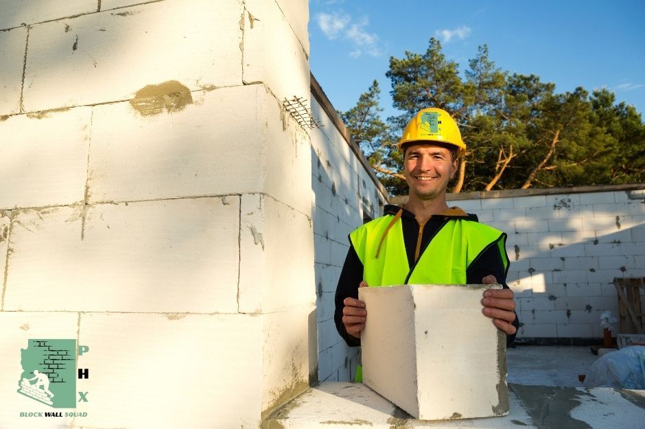 A block wall repair specialist wearing a bright yellow hard hat and high-visibility green safety vest is standing confidently. White concrete masonry units (CMU blocks) are neatly stacked and organized as part of a commercial repair project. He is working near E Whisper Rock Trail, and E Old Field Rd, Scottsdale, AZ.