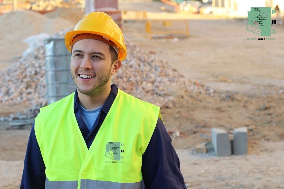 A block wall repair technician in orange hard hat and yellow safety vest smiles at a commercial cinder block retaining wall project, with gravel piles and concrete blocks behind him, near W 5th Ave, and S Sierra Vista Dr in Apache Junction, AZ.