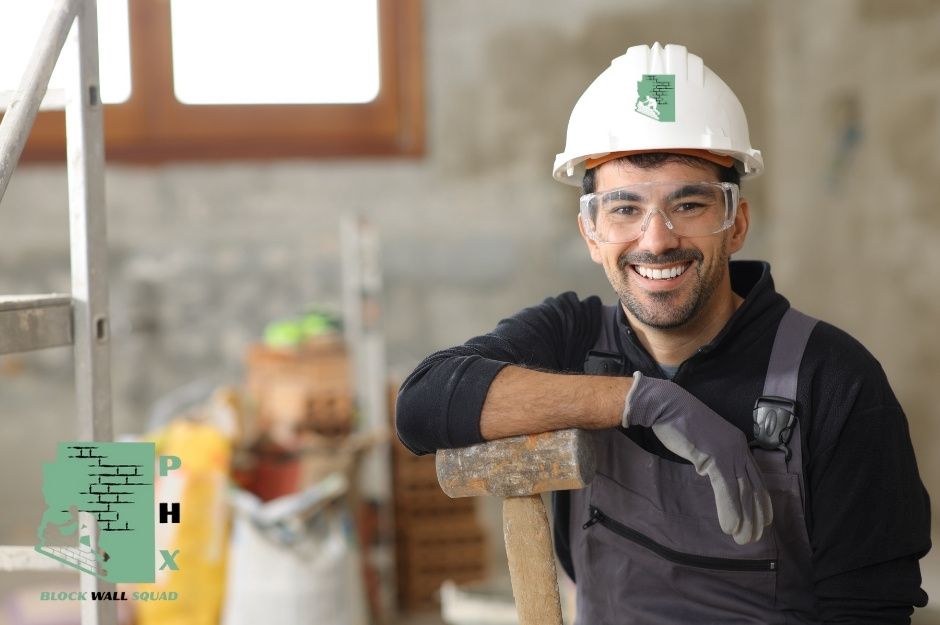 A smiling block wall repair technician in a white hard hat and dark work shirt poses with a hammer tool at a commercial construction site. The worker displays a confident, professional demeanor in the indoor construction environment near W Court St, and North Point Ridge Rd in Buckeye, AZ.
