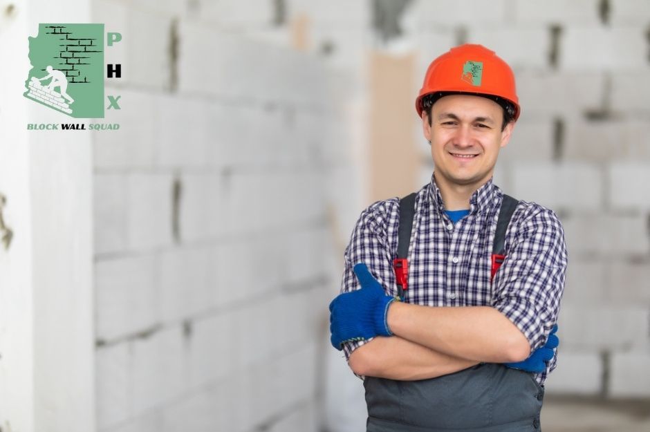 A confident block wall repair technician wearing an orange hard hat and blue work gloves stands with arms crossed in front of a white concrete block wall. The skilled worker appears to be ready to assess and repair a residential masonry work near W Azure Ln, and W Verbena Ln in Litchfield Park, AZ.