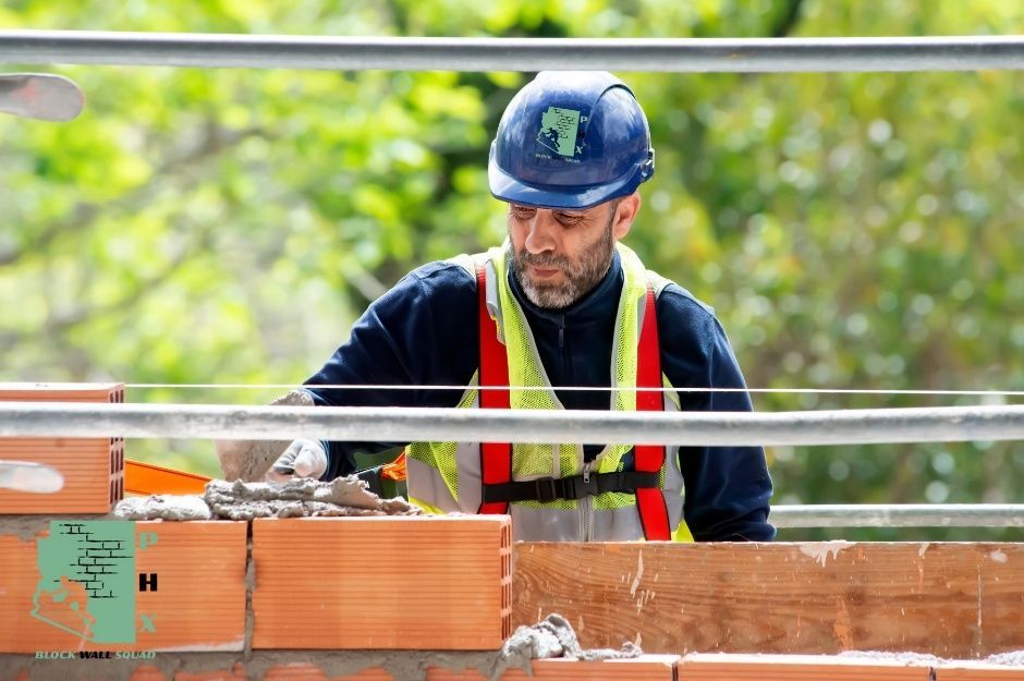 A block wall repair technician wearing a blue hard hat and safety vest works on a commercial masonry project, using a trowel to repair red brick blocks. The contractor focuses on precise mortar application near S Brentwood Dr, and S Howard Dr in Sun Lakes, AZ.