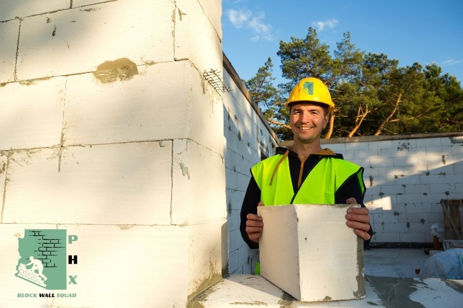 A smiling block wall repair technician wearing a yellow hard hat and bright green safety vest holds concrete blocks while working on a residential white block wall. The professional worker is positioned next to the masonry structure, ready to install blocks near N 88th Ave, and W Adams St in Tolleson, AZ.