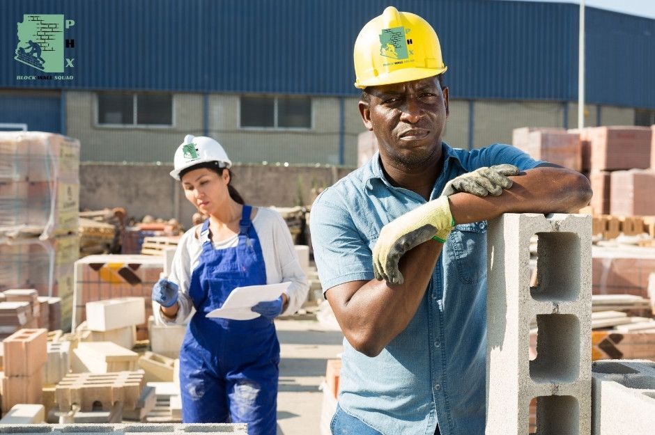 Block wall repair technicians work on a commercial masonry project, with the lead worker is in a yellow hard. A second technician in blue overalls and white hard hat assists in the background. Concrete blocks are stacked throughout the construction site at W Kennedy Ave, and N Main St in Coolidge, AZ.
