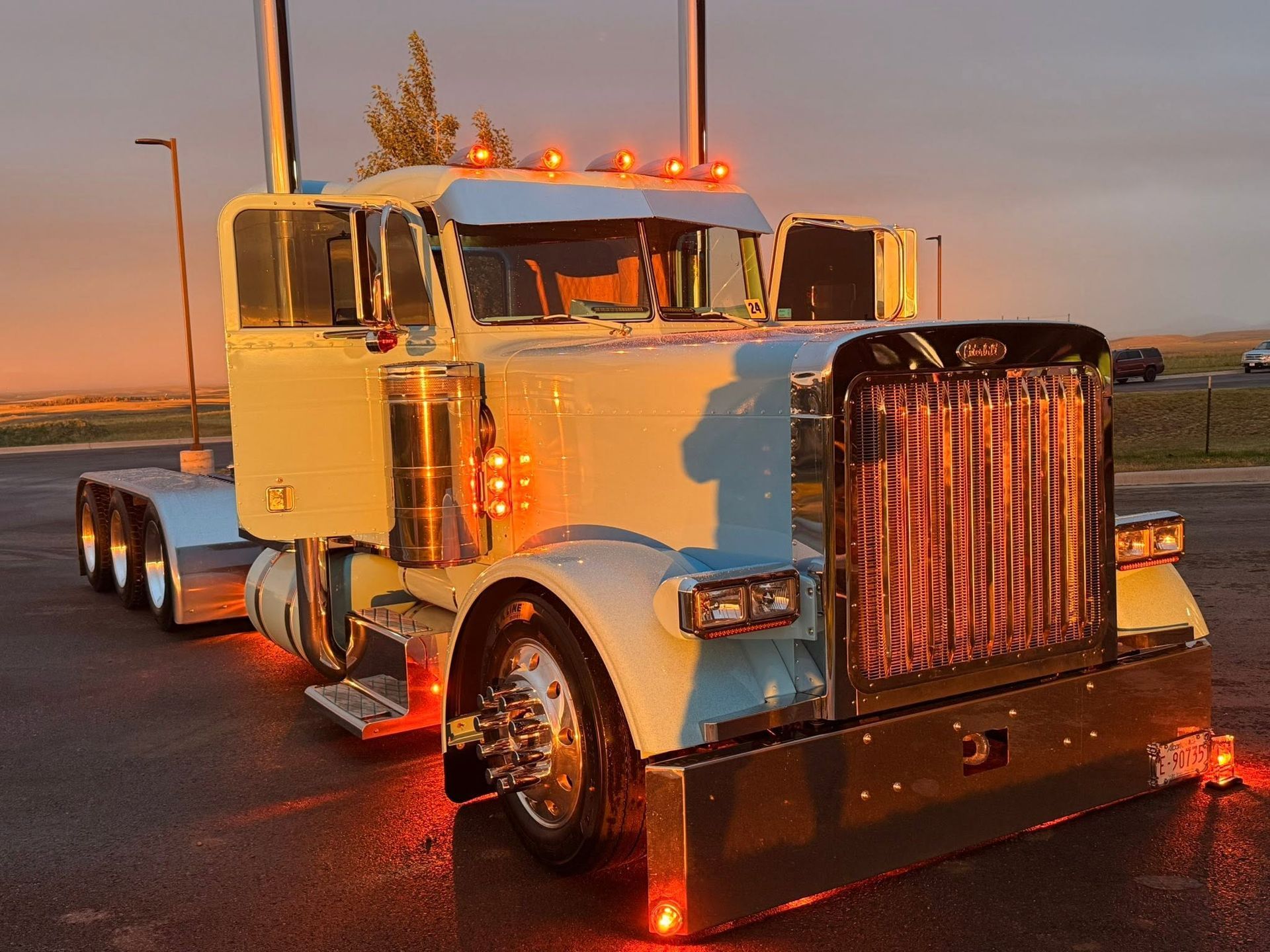 White semi truck with chrome grille and amber lights parked at sunset on a wet road