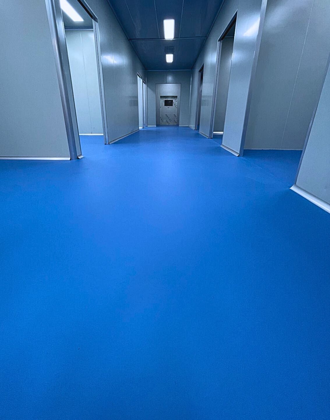 Blue-floored corridor with gray walls and doorways, brightly lit by overhead lights.