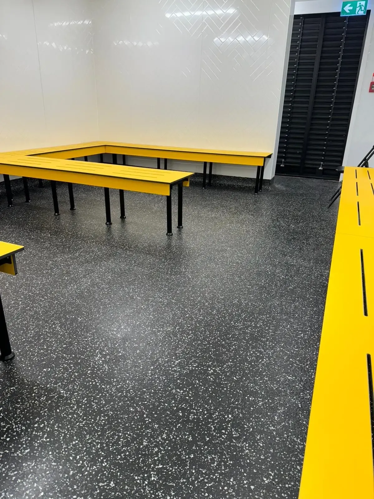 Yellow benches line a black-speckled floor in a brightly lit room with a white wall and a dark, slatted door.