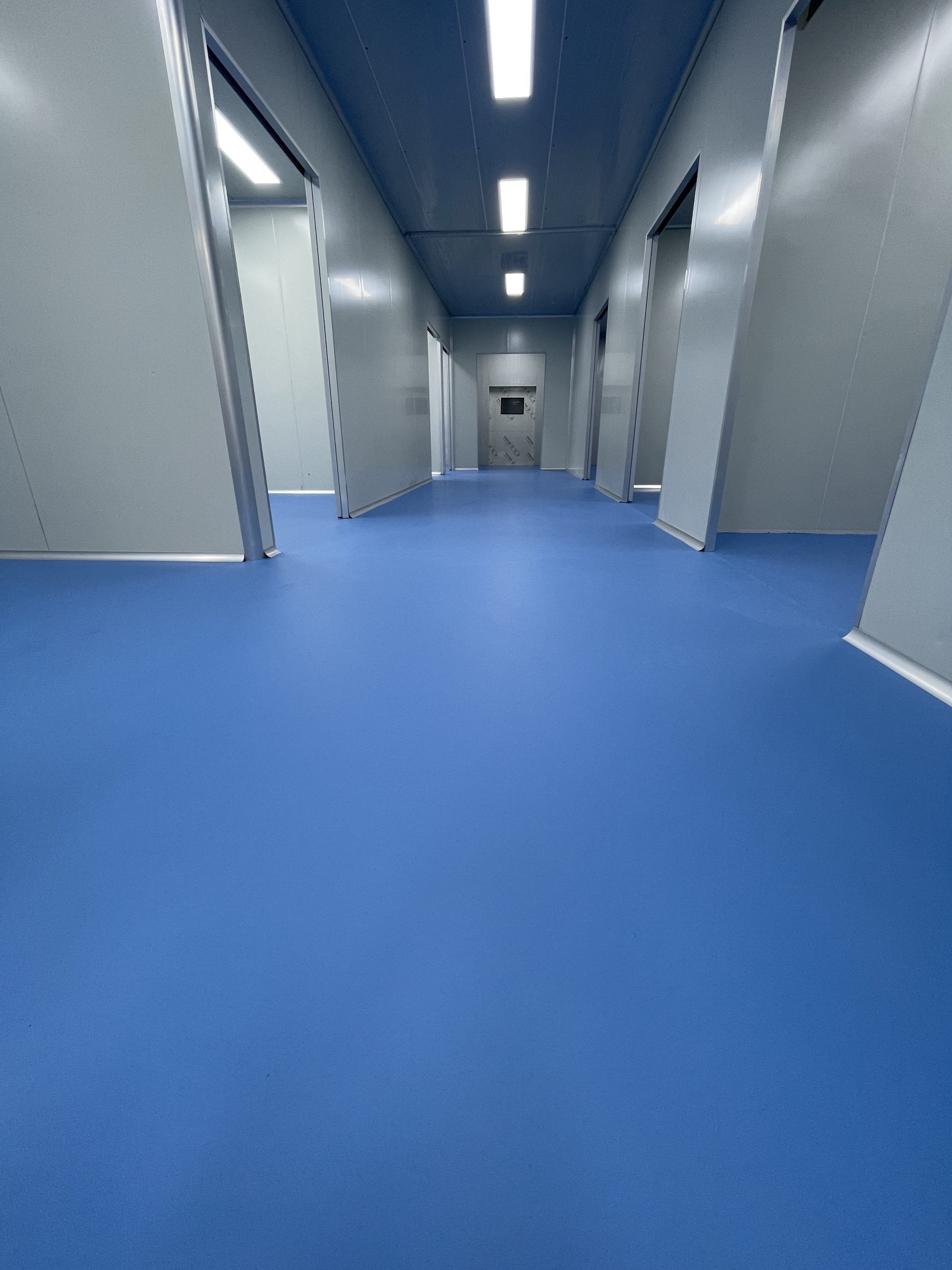 Long, blue-floored corridor with closed gray doors. Bright overhead lights.
