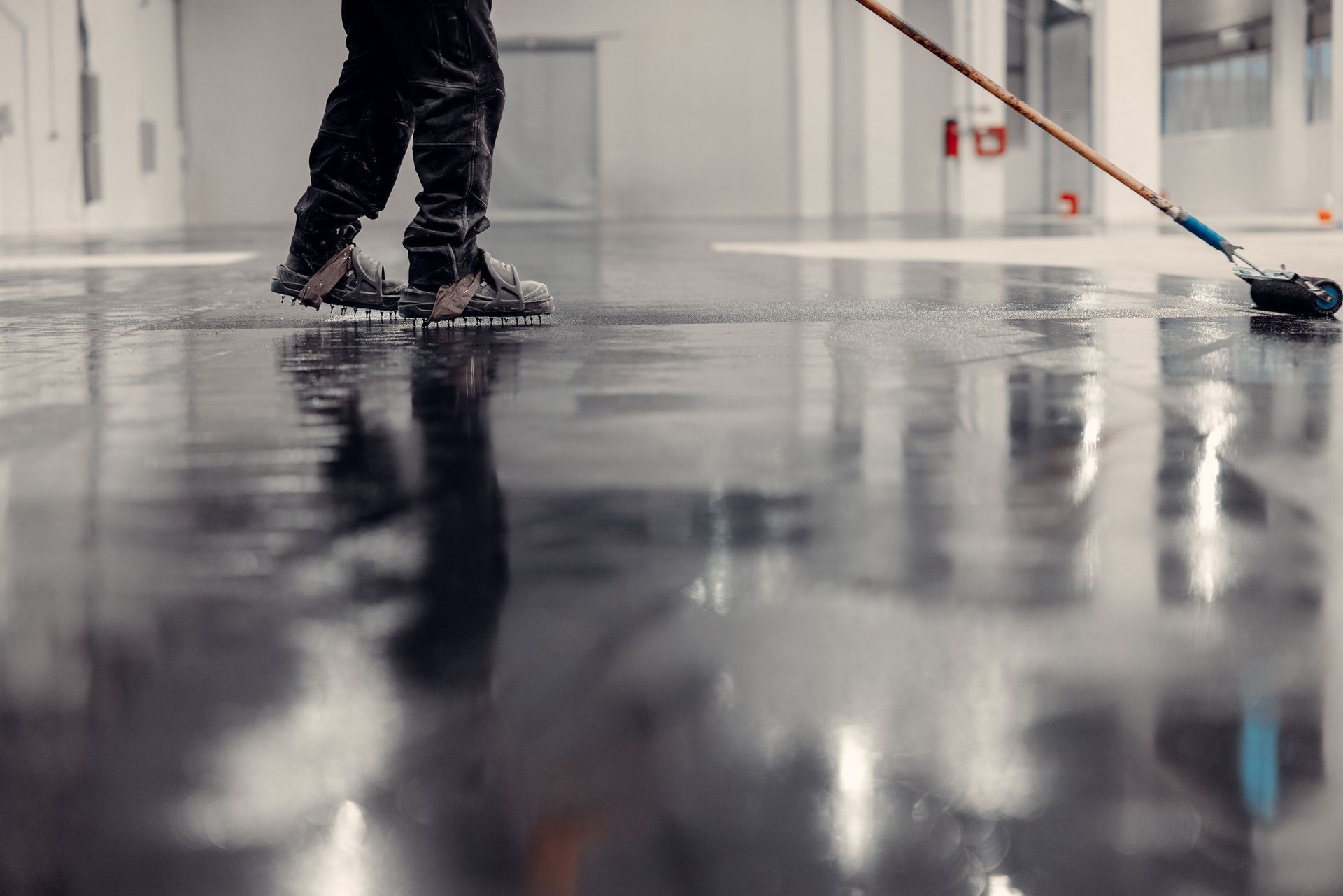 Person applying sealant to a dark, reflective concrete floor with a mop in a large, bright room.
