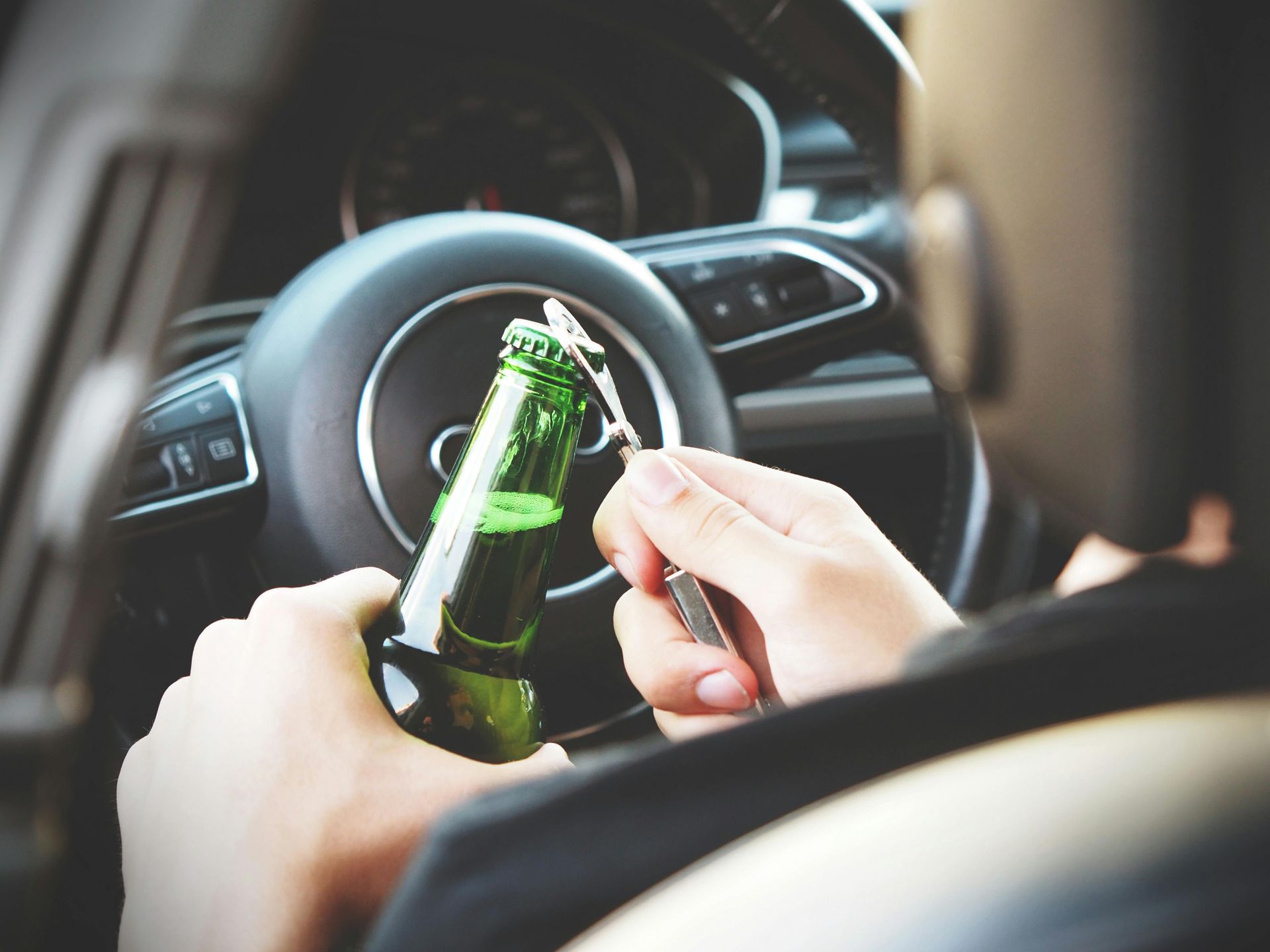 A person holds a green glass bottle and a bottle opener while sitting in the driver's seat of a car.