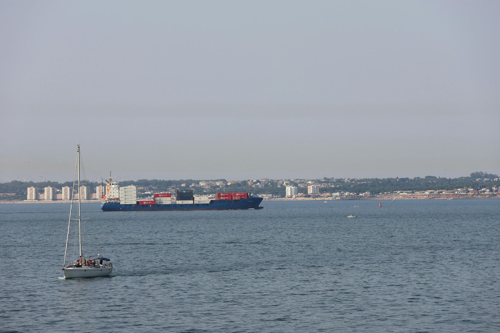 A sailboat glides on calm water in the foreground, with a large cargo ship sailing parallel to a distant, hazy coastline.