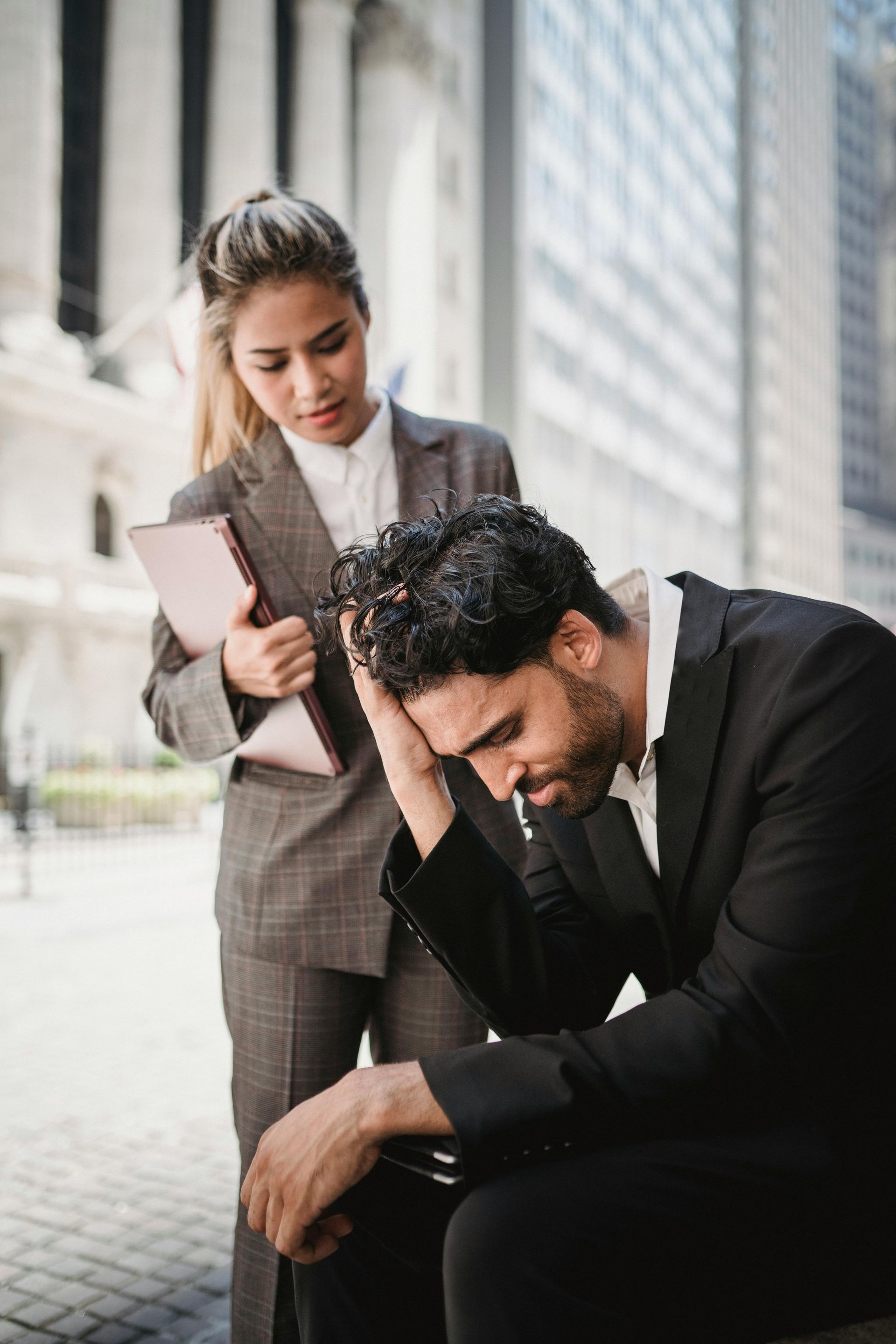 A woman in a grey suit holds a folder while looking down at a man in a black suit sitting with his head in his hands.
