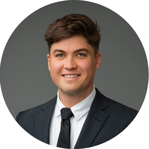 A person smiling at the camera wearing a dark suit, white collared shirt, and tie, against a solid gray circular background.
