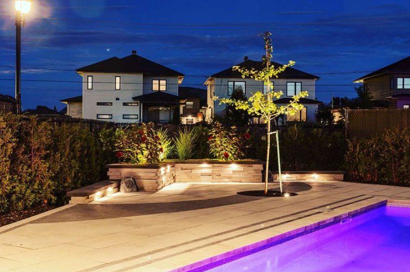 Une piscine est éclairée la nuit dans une cour arrière avec des maisons en arrière-plan.