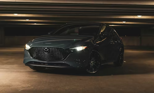 Dark gray sedan parked in a dim garage, front view with headlights on