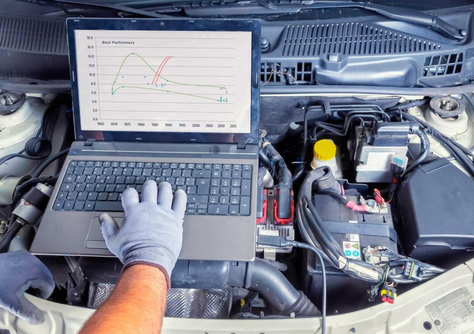 A man is using a laptop computer under the hood of a car.